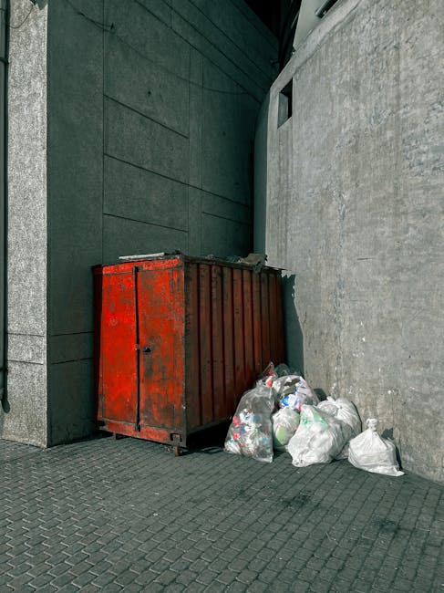 A large, rusted metal waste skip with a red finish, positioned against an exterior wall at the corner of a building. The skip's surface shows signs of weathering, with patches of corrosion and peeling paint. Surrounding the skip are several white plastic rubbish bags filled with waste, some of which are partially open, revealing mixed debris inside. The bags are placed directly on a paved sidewalk made of dark grey interlocking bricks. To the left, the building facade features smooth grey stone panels, while behind the skip, there is a dark green wall with a textured finish. To the right, a plain, coarse concrete wall extends upward, with a small rectangular opening near the top. The scene is lit by natural daylight, with no shadows or artificial lighting visible. This setup suggests an on-site rubbish collection area, possibly for private waste disposal, managed by local waste removal services such as Rubbish Removal Lambeth, highlighting alternative waste management methods outside typical council collection routines.