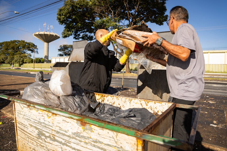 Two men are engaged in rubbish collection at an outdoor waste disposal point on a bright, sunny day. The man on the left, dressed in black with yellow gloves, is unloading a cardboard box filled with debris into a large, open-topped metal container that shows signs of rust and wear. The second man, wearing a light gray T-shirt and glasses, is assisting by holding the side of the box, helping to guide the contents into the container. Several black plastic rubbish bags and additional debris are already inside or nearby, some on a weathered, rust-streaked cart or trolley. In the background, there is a clear blue sky intersected by overhead power lines, along with a few leafy trees and a water tower, indicating an outdoor setting likely near a community or industrial area. The environment is well-lit by natural sunlight, emphasizing the textures of the waste materials and the metal surfaces, highlighting the practical and functional nature of waste management tasks performed by a private waste removal service like Rubbish Removal Lambeth.