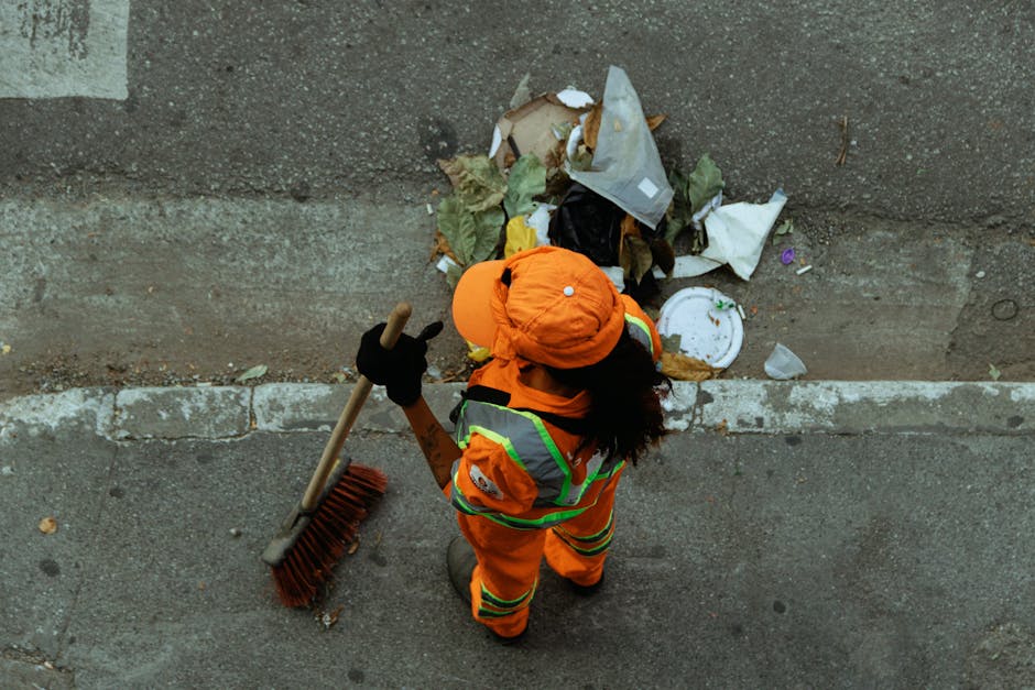 An individual dressed in high-visibility orange work clothing and a matching orange cap stands on a paved sidewalk next to a concrete wall. They are holding a broom with a wooden handle and red bristles, and appear to be in the process of clearing litter from the street. The person's back is partially turned to the camera, showing dark shoes and gloves. In front of them, there is a pile of assorted waste materials against the wall, including crumpled paper, plastic containers, and food wrappers, which are partially scattered on the pavement. The environment appears to be an urban street setting, with marked parking bays visible on the ground. The lighting is natural, suggesting daytime, and the scene emphasizes the activity of waste collection, relevant to private rubbish removal or on-site clearance services provided by companies like Rubbish Removal Lambeth.
