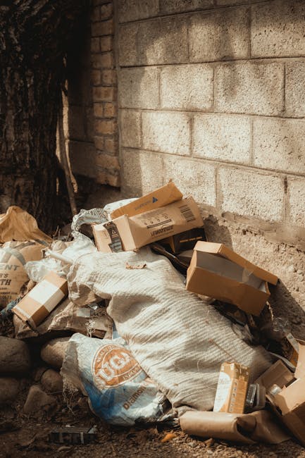 A pile of discarded cardboard boxes, some flattened and others partially intact, along with crumpled paper and plastic packaging, is seen leaning against a rough-textured brick wall outdoors. The boxes vary in size, with some showing printed labels and barcodes, while the paper packaging appears torn and soiled. A large woven sack, possibly used for waste collection, is partially covering the boxes and has a rough, fibrous surface with some dirt marks. Surrounding the pile are small rocks and soil, indicating an outdoor environment, possibly near a building or garage. The brick wall behind the waste exhibits a weathered appearance with visible mortar joints, and the overall scene suggests an informal collection of domestic rubbish awaiting removal or disposal. The lighting appears natural, highlighting the textures and colors of the materials, consistent with an area for private or independent waste handling that might be preferred for quick on-site clearance or alternative rubbish removal services implemented by Rubbish Removal Lambeth.