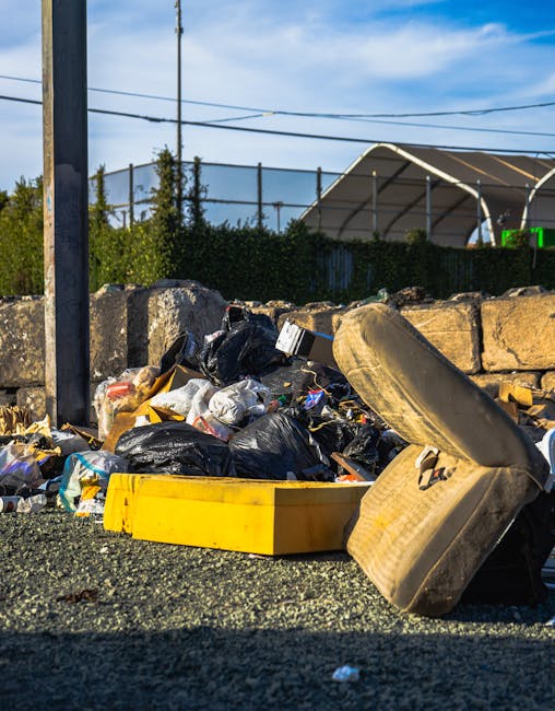 An outdoor scene showing a pile of mixed household rubbish including black plastic bags, newspapers, and small discarded items, spread across a gravel surface beside a low stone wall. Prominent in the foreground is a pair of beige upholstered chairs stacked haphazardly, with visible fabric texture and minor staining. To the left, a yellow plastic container lies on its side, partially filled with waste. In the background, a metal pole supports electrical wires overhead, and a fencing structure surrounds a tennis court with a curved, partially open roof visible beyond the fence. Green foliage and a clear blue sky with light clouds are visible behind the scene, indicating daylight and outdoor conditions. The image context suggests a location where private waste disposal or on-site waste handling occurs, aligning with alternative rubbish removal solutions such as those offered by Rubbish Removal Lambeth.
