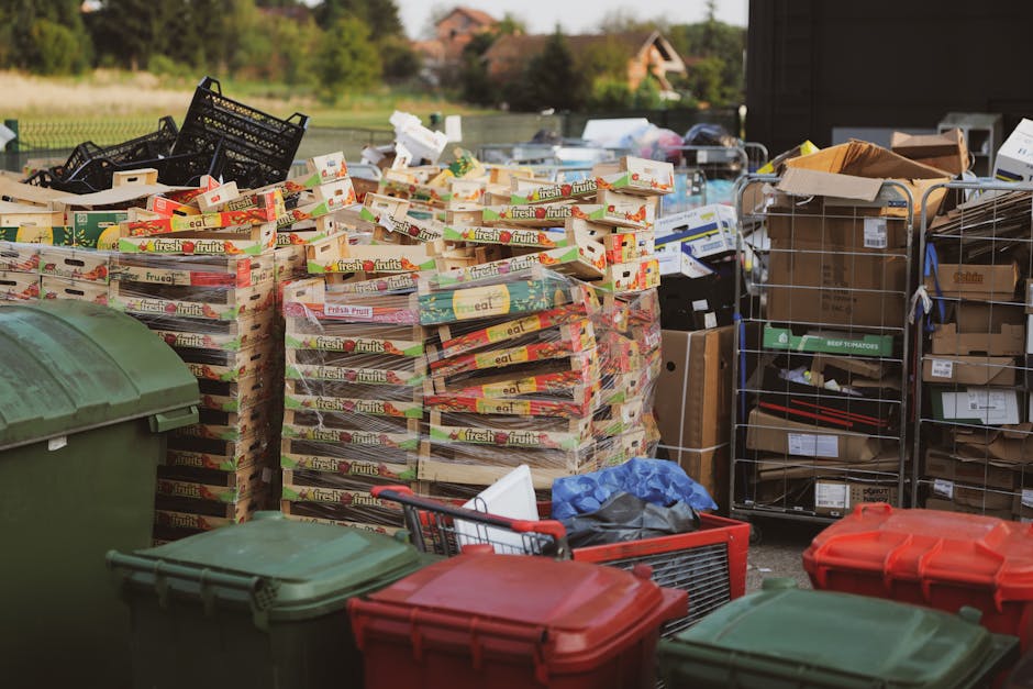 A pile of discarded cardboard boxes, some flattened and others partially intact, along with crumpled paper and plastic packaging, is seen leaning against a rough-textured brick wall outdoors. The boxes vary in size, with some showing printed labels and barcodes, while the paper packaging appears torn and soiled. A large woven sack, possibly used for waste collection, is partially covering the boxes and has a rough, fibrous surface with some dirt marks. Surrounding the pile are small rocks and soil, indicating an outdoor environment, possibly near a building or garage. The brick wall behind the waste exhibits a weathered appearance with visible mortar joints, and the overall scene suggests an informal collection of domestic rubbish awaiting removal or disposal. The lighting appears natural, highlighting the textures and colors of the materials, consistent with an area for private or independent waste handling that might be preferred for quick on-site clearance or alternative rubbish removal services implemented by Rubbish Removal Lambeth.