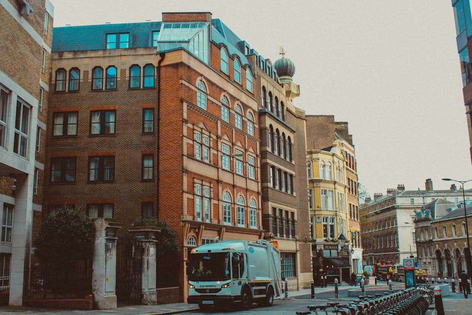 The image depicts a lively urban street scene featuring a row of historic-style buildings with brick facades, arched windows, and decorative architectural details. In the foreground, a light blue rubbish collection van is parked on the pavement near the curb, positioned on the right side of the image. The van has a clean, smooth finish with a compact design suitable for urban waste collection. To the left of the van, there is a small section of a gated entrance with metal fencing and stone pillars, partially obscured by trees and shrubbery. Surrounding the buildings are various elements of city life, including bicycles lined up along a rack in the lower right corner and streetlamps overhead, which provide ambient lighting. The scene is set during daylight hours with overcast weather, giving the environment a neutral, professional atmosphere relevant to private rubbish removal and on-site clearance services. The overall setting emphasizes a typical city street where independent waste collection services like Rubbish Removal Lambeth operate to manage waste outside local authority collection areas.