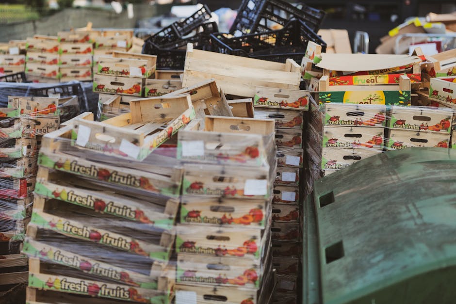 A large outdoor collection of discarded fruit boxes made of light-colored wood and black plastic crates, stacked irregularly across a paved surface. The wooden boxes feature colorful printed labels with images of fresh fruits and the words 'fresh fruits,' with some displaying visible white labels or tags attached. The black plastic crates are positioned at the back, leaning against or stacked on top of the wooden boxes, and show signs of wear and discoloration. To the right, there is a partially visible greenish container or structure, possibly a recycling bin or waste container, with a textured surface and a slightly weathered appearance. The scene appears to be an outdoor area designated for rubbish collection or disposal, typical of private waste removal services, with natural lighting and no human activity visible. Rubbish Removal Lambeth might be involved in managing this type of waste segregation or collection of fruit packaging materials.