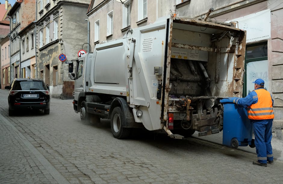 The image depicts a lively urban street scene featuring a row of historic-style buildings with brick facades, arched windows, and decorative architectural details. In the foreground, a light blue rubbish collection van is parked on the pavement near the curb, positioned on the right side of the image. The van has a clean, smooth finish with a compact design suitable for urban waste collection. To the left of the van, there is a small section of a gated entrance with metal fencing and stone pillars, partially obscured by trees and shrubbery. Surrounding the buildings are various elements of city life, including bicycles lined up along a rack in the lower right corner and streetlamps overhead, which provide ambient lighting. The scene is set during daylight hours with overcast weather, giving the environment a neutral, professional atmosphere relevant to private rubbish removal and on-site clearance services. The overall setting emphasizes a typical city street where independent waste collection services like Rubbish Removal Lambeth operate to manage waste outside local authority collection areas.
