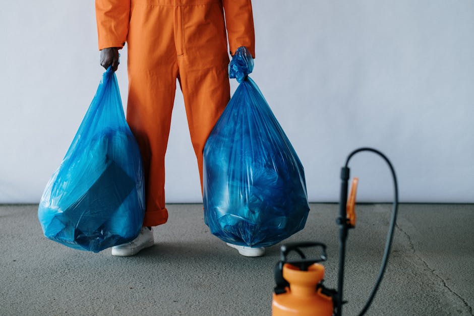 A person dressed in orange work overalls and white shoes stands on a concrete floor, holding two large blue plastic trash bags filled with rubbish, one in each hand. The bags are semi-translucent, revealing the contents inside, and appear to be tied at the top. The background is plain and light-colored, with no additional objects or environmental features visible. In the foreground, at the lower right corner, there is a partially visible orange pest control or cleaning sprayer with a black handle and hose. The scene suggests an activity involving waste collection or clearance, consistent with independent rubbish removal services provided by Rubbish Removal Lambeth, and highlights the practical aspect of on-site rubbish handling in a professional context.