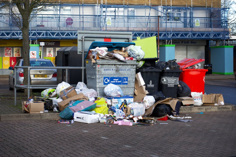 A large collection of overflowing waste bins and a variety of rubbish scattered on a paved urban street, including black, red, and grey bin bags, flattened cardboard boxes, paper and plastic packaging, and discarded miscellaneous items. The bins are positioned in front of a commercial building with storefronts, including a fish bar, and a blue metal construction scaffold partially covering the upper part of the building. A small parked car with a yellow license plate is visible to the left of the waste collection area. The scene is outdoors, with a tree on the left and a low metal safety barrier separating the waste from the pedestrian walkway. The overall environment appears cluttered, highlighting issues related to rubbish disposal and waste management, which could be addressed by independent waste collection services like Rubbish Removal Lambeth, as part of alternative rubbish handling options in urban areas.
