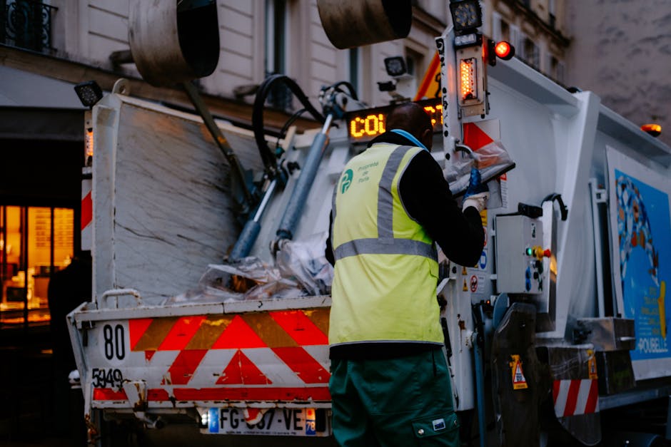 A worker wearing a high-visibility yellow vest and dark clothing operates a waste collection vehicle on an urban street during dusk. The vehicle is equipped with mechanical arms and digital display screens, and is actively engaged in the process of collecting waste materials from an open-top hopper. The side of the vehicle features red and white reflective markings and a blue logo, indicating it is used for professional rubbish removal services. In the background, a building with illuminated windows provides ambient lighting, casting a warm glow in contrast to the cooler, evening natural light. The waste materials inside the hopper appear to be a mix of paper, plastic, and other household items, piled loosely with some visible debris on top. The scene depicts an independent collection activity typical of private rubbish disposal, supporting standard waste management practices for urban tradespeople or local residents, aligning with professional rubbish removal standards. The focus on the machinery, waste contents, and environment emphasizes operational details relevant to waste collection services provided by companies like Rubbish Removal Lambeth.