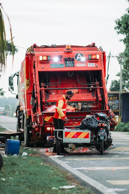 A waste collection technician is operating a large red rubbish truck on the side of a street, positioned on the curb with its rear compartment open for waste disposal. The technician, wearing a fluorescent yellow safety vest over a red long-sleeved shirt, is seen loading or managing garbage into the truck's designated compartment, which features hydraulic lifting mechanisms and metal surfaces with signs of wear. Nearby, a black plastic bag filled with waste is placed on a small trailer or cart attached to the truck. The scene takes place during daylight hours with overcast skies, and the surrounding environment includes a sidewalk, some greenery, and a background with trees, power lines, and a few commercial signs, indicative of an urban or suburban setting. This image reflects an on-site waste collection process that could relate to independent or private rubbish removal services like those offered by Rubbish Removal Lambeth, supporting alternative waste handling methods outside local authority collections.