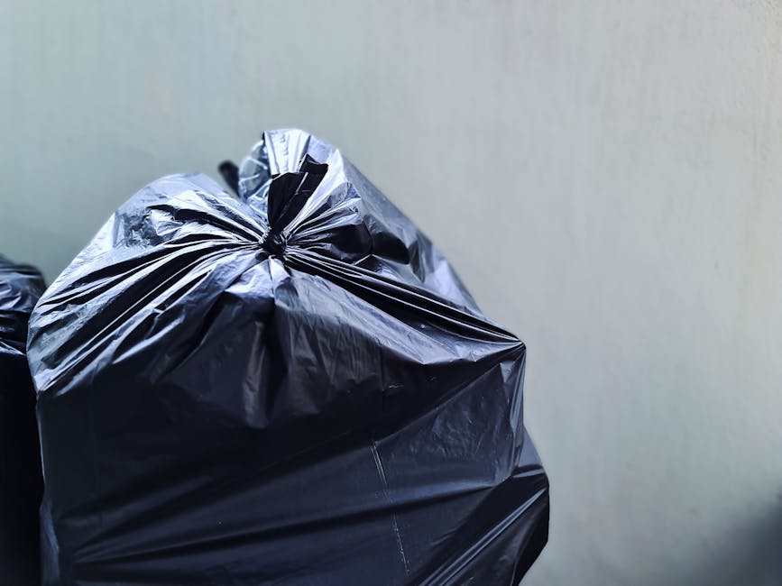 A close-up view of several tightly tied black plastic rubbish bags placed against a plain, light-colored wall. The bags appear to be made of glossy, durable plastic material, with some showing slight wrinkles and reflections from ambient light. The bags are situated on a flat surface, likely outdoors or on a pavement, with one bag partially overlapping the others. The scene suggests a waste collection or disposal scenario typical of private rubbish removal services, such as those offered by Rubbish Removal Lambeth, involving the on-site clearing and loading of household or commercial waste. The overall composition emphasizes practical waste containment, with the focus on the full, sealed rubbish bags prepared for collection, aligning with standard practices for independent waste disposal in urban environments.