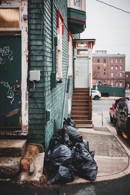 Two large black rubbish bags made of thick plastic material, with visible creases and folds, are placed on the pavement next to a curb. The bags appear to be partially filled and are leaning slightly, with one positioned more upright and the other resting at an angle. Behind the bags, there is a wooden fence painted in dark colors, with vertical slats spaced closely together, and dense foliage with leafy branches extending above and around the fence. The scene is set outdoors on a street or alleyway, and the lighting suggests overcast or shaded conditions, contributing to a subdued, neutral tone. The presence of these bags indicates an active rubbish removal process, possibly part of a private waste collection service such as Rubbish Removal Lambeth, highlighting options beyond municipal disposal methods. Overall, the scene exemplifies typical on-site rubbish clearance, with emphasis on the practical handling and disposal of household or garden waste in an urban environment.