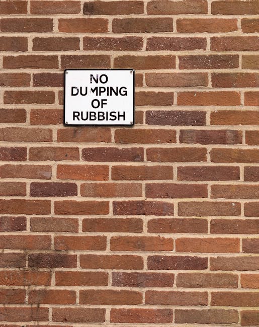 A rectangular metal sign mounted on a red-brown brick wall, displaying the message 'NO DUMPING OF RUBBISH' in black capital letters on a white background. The sign is positioned approximately one-quarter from the top of the image and is centered horizontally. The brick wall behind the sign features a traditional running bond pattern, with alternating rows of full and half bricks, varying in shades of red, brown, and orange, with light-colored mortar joints between them. The lighting appears natural, casting soft shadows at the bottom of the sign and highlighting the texture of the bricks. The sign serves as an official notice for private property or local waste management, indicating restrictions on rubbish disposal in the vicinity, which aligns with services provided by rubbish removal specialists like Rubbish Removal Lambeth.