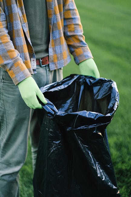 A person standing outdoors on a grassy area, wearing a plaid shirt with mustard yellow, gray, and blue tones over a dark green top, paired with gray trousers and a brown belt. They are holding open a black plastic rubbish bag, lined with a silver inner layer, with both hands wearing bright green disposable gloves. The background features well-maintained green grass, suggesting a garden or park environment, and the scene appears to be related to rubbish collection or waste disposal, consistent with private waste management services. The image focuses on the individual's torso and hands, with no visible face, and emphasizes the action of handling waste in an organized, professional manner, indicative of rubbish removal activities.