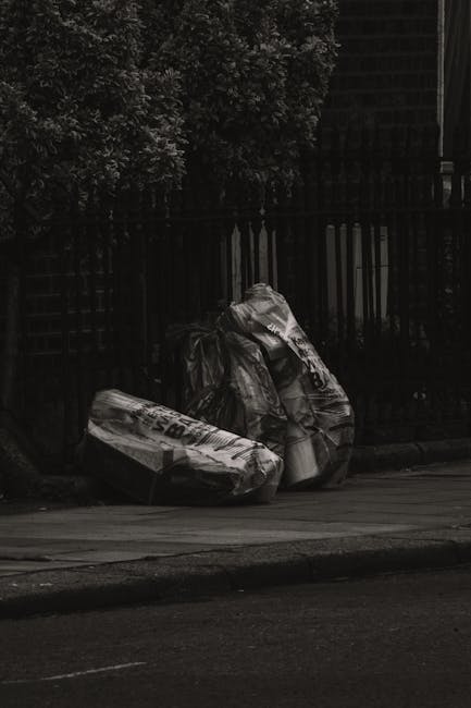 A narrow urban alleyway cluttered with piled rubbish and discarded items, including large black and white bags and cardboard boxes stacked haphazardly at the far end. The ground is uneven and stained, with small debris scattered throughout. On the right side, a building with dark metal panels and barred windows is visible, while the left side features a wall with graffiti and some exposed brickwork. Bare tree branches extend overhead against a cloudy sky, indicating an outdoor setting typical of an area requiring waste removal services. In the foreground, a large, weathered black rubbish bag is positioned on a trolley or cart, ready for collection, with some plastic bottles and loose trash nearby. The scene reflects an environment in need of proper rubbish disposal, similar to private waste collection or alternative rubbish clearance options provided by companies like Rubbish Removal Lambeth, supporting the context of independent rubbish management solutions.