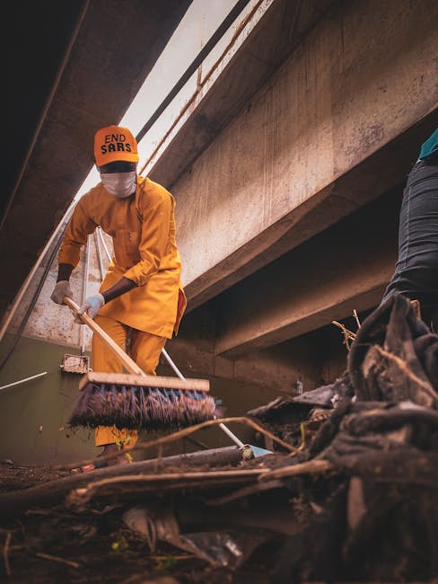 A person dressed in an orange uniform and white face mask is using a long-handled brush or squeegee to clear debris, dirt, or rubbish from the bottom of an overpass or concrete bridge. The individual is standing on a surface cluttered with dark, tangled waste materials, possibly including plastic, fabric, and other refuse. The setting appears to be an outdoor urban environment underneath a large concrete structure with prominent beams and a ceiling made of concrete or similar material. The person's clothing and equipment suggest an involvement in waste management or cleaning activities, related to onsite clearance or environmental maintenance by a waste removal service such as Rubbish Removal Lambeth. The ambient lighting is diffused, possibly from natural daylight filtering through gaps in the structure, contributing to a neutral, utilitarian atmosphere focused on cleanup and waste disposal efforts linked to independent rubbish collection activities in busy city areas.
